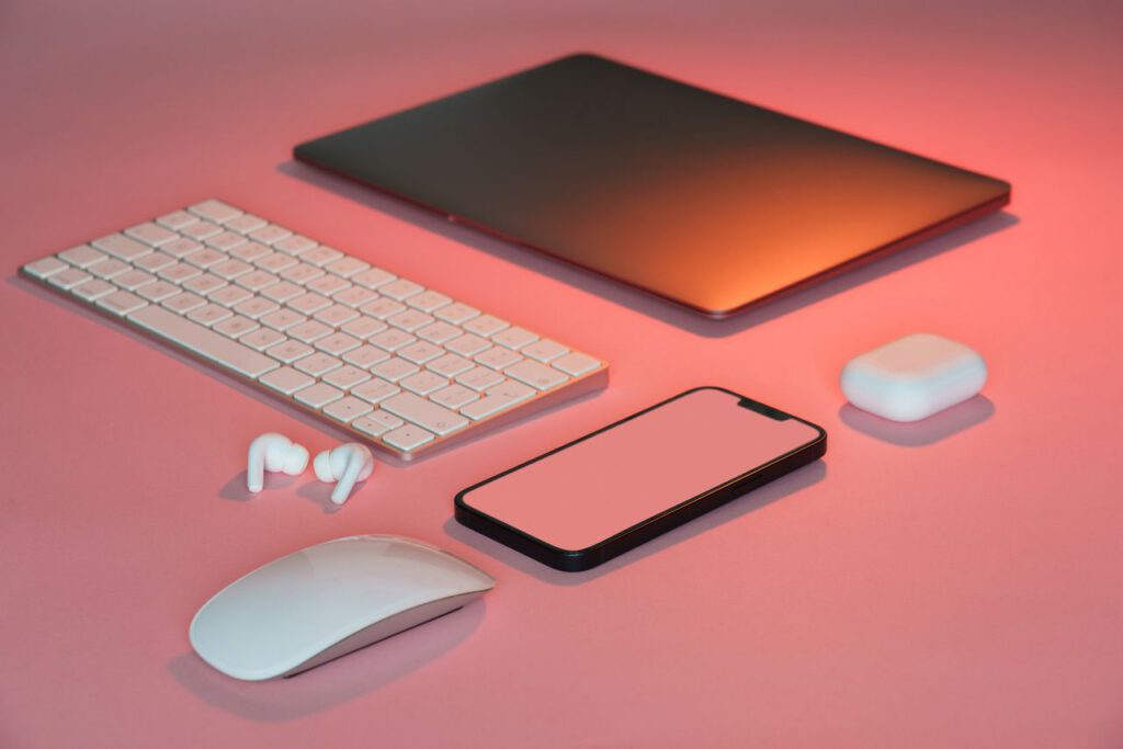 Flat lay of modern tech gadgets on a pink surface, featuring a keyboard, mouse, laptop, smartphone, earbuds, and case.